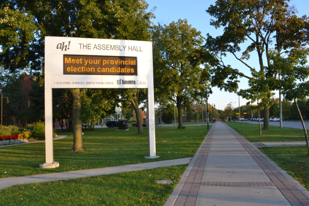 Ein Schild auf dem Boden mit der Aufschrift "Die Versammlungshalle - Treffen Sie Ihre Landtagswahlkandidaten", umgeben von Gras, einem Weg, Bäumen, blühenden Pflanzen, Straßenlaternen, Fahrzeugen, einem Gebäude mit Fenstern und einem bewölkten Himmel.