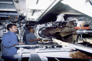 Drei Männer in einer Fabrik inspizieren eine große Maschine, umgeben von verschiedenen Werkzeugen und Geräten.