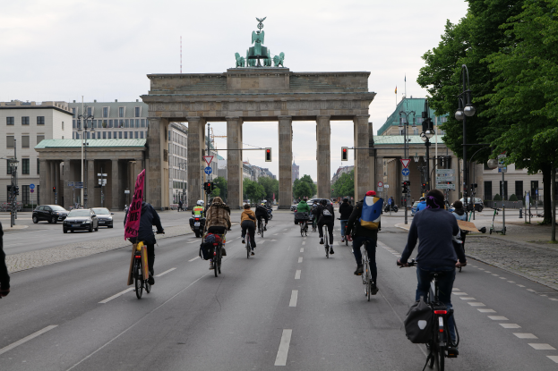 Eine Gruppe von Menschen, die auf Fahrrädern die Straße vor dem Brandenburger Tor in Berlin, Deutschland, entlangfahren, mit Bäumen, Laternen, Ampeln und Gebäuden mit Fenstern im Hintergrund, unter einem bewölkten Himmel.
