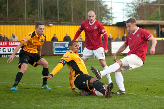Spieler in blauen und roten Uniformen konkurrieren in einem Spiel auf einem grünen Feld, mit einem Ball und einem Baum sowie Zuschauern, die außerhalb des Feldes stehen und jubeln.