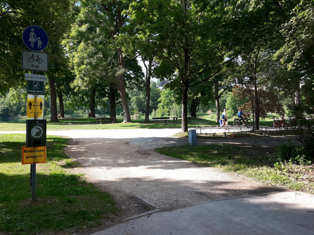 Ein gepflasterter Parkweg, der von Bäumen und Gras gesäumt ist, mit Schildern an einem Pfahl auf der linken Seite und Radfahrern im Hintergrund.
