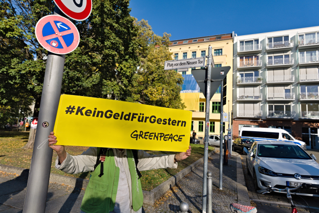 Eine Person mit einer Tasche und einem "Greenpeace"-Schild vor einem Gebäude mit Fahrzeugen, einem Fahrrad und Schildern in der Nähe, mit Bäumen und einem klaren blauen Himmel im Hintergrund.