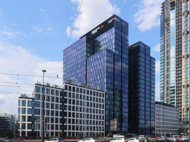 Zentrale der Deutschen Bank in Frankfurt, Deutschland, ein großes mehrstöckiges Gebäude mit vielen Fenstern, umgeben von städtischer Infrastruktur einschließlich Straßenlaternen, elektrischen Kabeln, Fahrzeugen, Grünflächen und einem bewölkten Himmel.