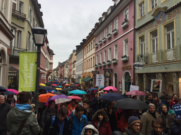 Große Gruppe von Menschen, die mit Schirmen die Straße entlanggehen, einige tragen Taschen und halten Schilder, während einer Klimademonstration in Deutschland, mit Gebäuden, die die Straße säumen und einem Laternenpfahl im Vordergrund.