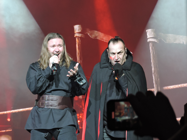 Ozzy Osbourne und eine weitere Person in schwarzen Kleidern auf der Bühne der O2 Arena in London, mit einer Person im Vordergrund, die ein Mobiltelefon hält, und Seilen, Lichtern im Hintergrund.