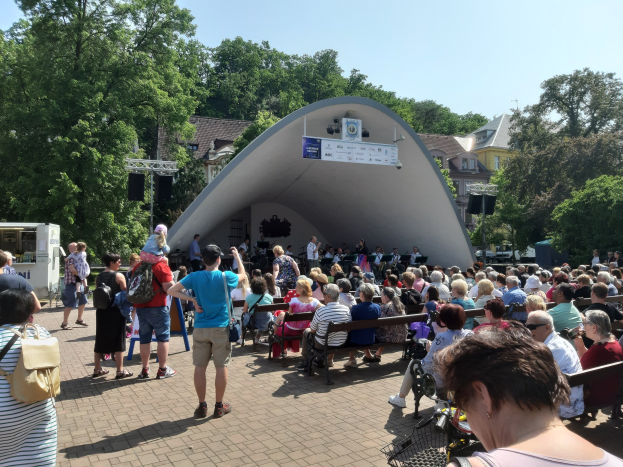Eine Menschenmenge sitzt auf Bänken und steht vor einer Bühne bei einem Open-Air-Konzert im Sommer, mit einem Kameramann, einem Mikrofonständer, einer Fahne, Bäumen, Gebäuden und einem bewölkten Himmel.