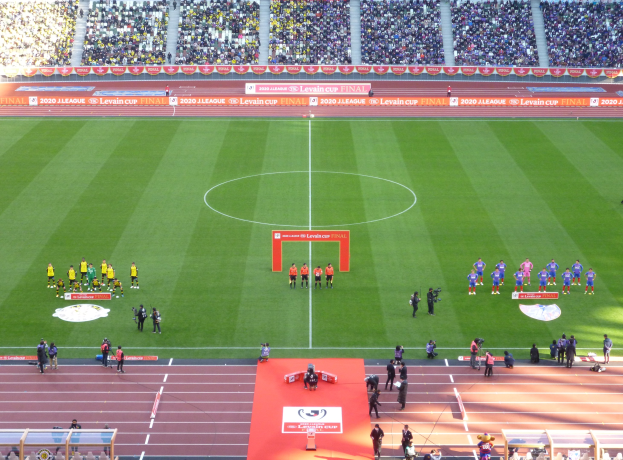 Eine Gruppe von Menschen steht auf einem grünen Fußballfeld in einem Stadion voller Zuschauer, mit Werbetafeln für das 2020 Levin Cup-Finale im Hintergrund.