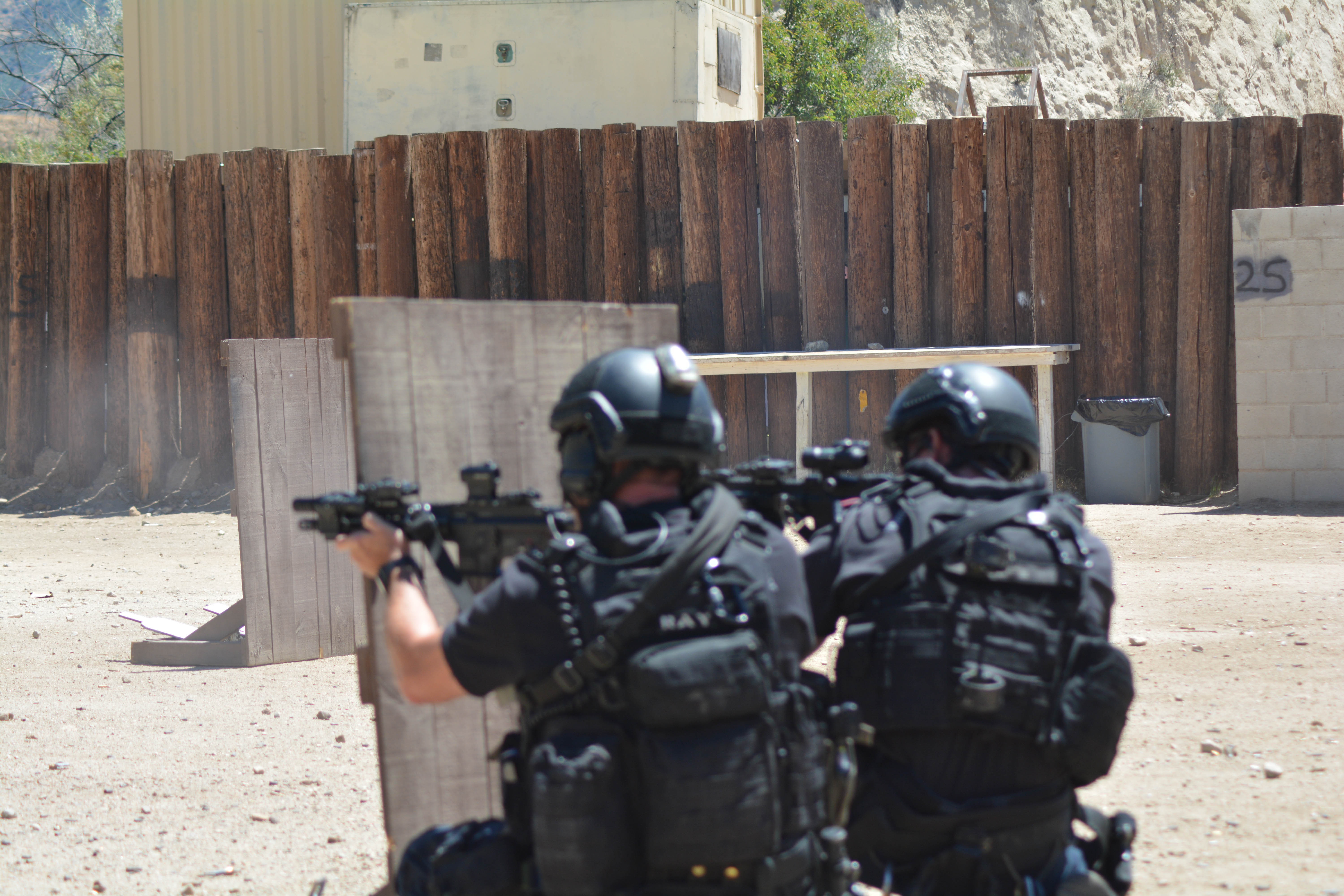 Zwei Polizisten in schwarzen Uniformen und Helmen, die Waffen in den Händen halten, stehen vor einem Holzzaun mit einer Wand zur Rechten und einem Gebäude im Hintergrund, mit Bäumen und einem Hügel in der Ferne.