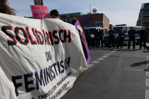 Eine Gruppe von Menschen marschiert auf einer Straße und hält ein 'Solidarität und Feminismus'-Schild, mit parkenden Fahrzeugen, Gebäuden, einer Schüsselantenne und einem klaren blauen Himmel im Hintergrund.