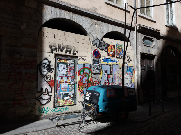 Ein blaues und schwarzes Fahrzeug, das auf der Straße neben einem Gebäude mit buntem Graffiti geparkt ist, Fenster und Türen, sowie ein Mast und Kabel, die entlang der Straße verlaufen.