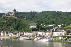 Ein malerischer Blick auf den Rhein in Deutschland mit einer Burg auf einem Hügel im Hintergrund, Booten auf dem Fluss und Fahrzeugen auf einer nahen Straße sowie einem bewölkten Himmel.