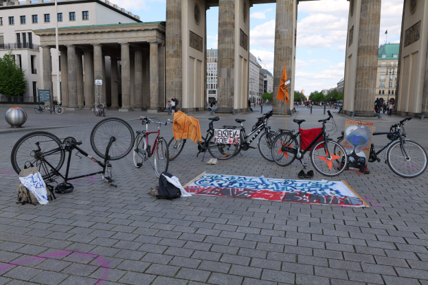 Eine Gruppe von Fahrrädern vor dem Brandenburgertor in Berlin, Deutschland, mit Taschen, Gegenständen und einer Fahne mit Text, Gebäuden, Bäumen, Polen, Flaggen und einem klaren blauen Himmel im Hintergrund.