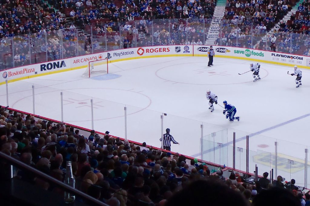 Vier Männer spielen Hockey auf einem Eisplatz, mit zwei in der Nähe des Netzes und des Zaunes stehend, Zuschauern auf Stühlen im Vorder- und Hintergrund und einer Treppe in der Ferne sichtbar.
