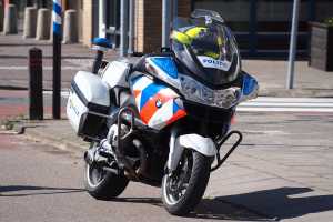 Ein Polizei-Motorrad am Straßenrand mit einem Helm darauf, mit Pfählen, Pflanzen und einem Gebäude im Hintergrund.