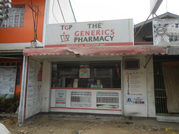 Ein Apothekenschild mit der Aufschrift "The Generics Pharmacy" vor einem Gebäude mit umliegenden städtischen Elementen unter einem klaren Himmel.