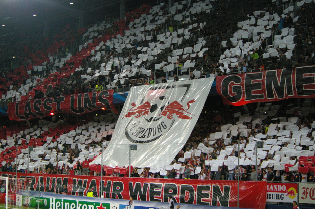 Ein volles Fußballstadion mit Sitz- und Stehplätzen, Bannern, einem Torpfosten und Flutlicht.