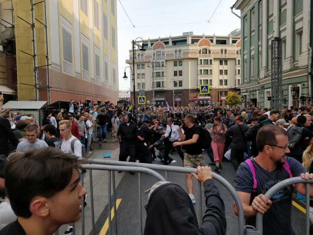 Eine große Menschenmenge steht vor einer Metallabsperrung auf einer Straße, umgeben von Gebäuden, Laternen, Schildern, Bäumen und einem klaren Himmel, wobei einige Individuals mit Kameras zu sehen sind.