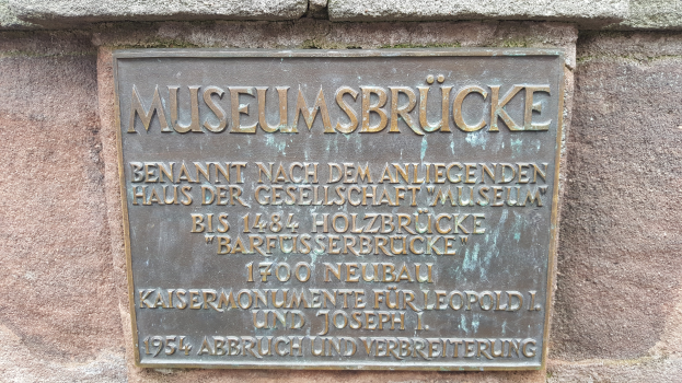Eine dunkle Plakette auf einem Steingebäude trägt die Inschrift "Museumsbrucke" in fetter Schrift.