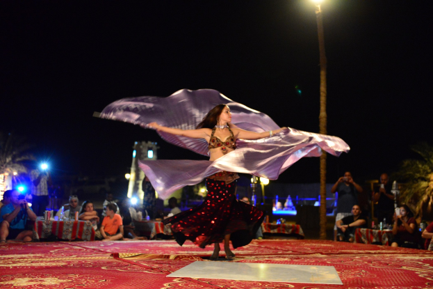 Eine Frau in einem Bauchtanzkostüm tanzt auf einem roten Teppich, umgeben von einer Zuschauermenge mit Kameras, mit Bäumen, Lichtern und einer Stange im Hintergrund.