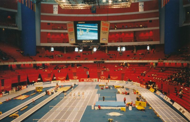 Ein großes Hallenstadion voller Zuschauer bei einer Leichtathletikveranstaltung mit Sitzplätzen, Stehplätzen, Texttafeln, Lichtern und einem Bildschirm oben.