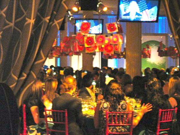 Eine Gruppe von Menschen an Tischen mit Essen, Fernsehern, Lichtern, hängenden Papieren und Vorhängen in einer Art Party-Setting.