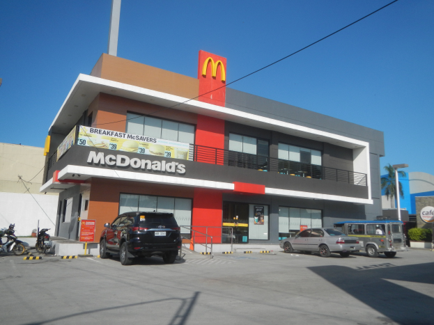 Ein McDonald's-Restaurant mit parkenden Autos davor, umgeben von Gebäuden, Straßenlaternen, Strommasten, Stromkabeln, Fahrzeugen auf der Straße, Infotafeln, Büschen, Bäumen und einem klaren blauen Himmel.
