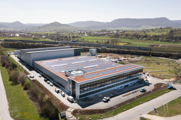 Eine Luftaufnahme eines großen Fabrikgebäudes mit Solarmodulen auf dem Dach, umgeben von Fahrzeugen, Bäumen und Masten, mit Hügeln und einem klaren blauen Himmel im Hintergrund.