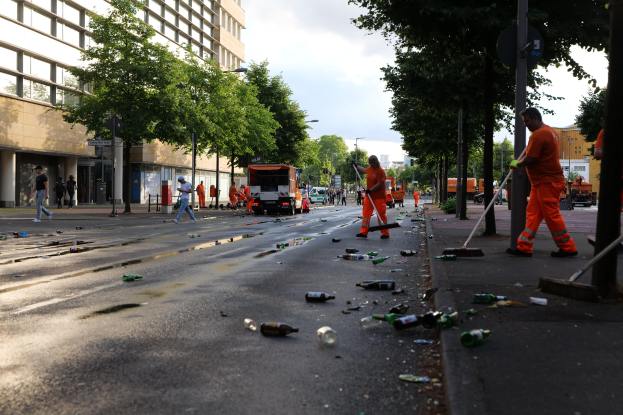 Eine Gruppe von Menschen in orangefarbenen Uniformen sammelt Müll von einer Straße, die mit Flaschen und Schutt übersät ist, mit Bäumen, Pfählen und Fahrzeugen im Hintergrund.