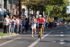 Zwei Personen mit Helmen und Brillen fahren auf Fahrrädern eine von Bäumen gesäumte Straße entlang, während eine Gruppe von Menschen mit Kameras auf dem Gehweg zur Linken steht, ein Band an einem Pfahl befestigt, der Hintergrund unscharf, um Bewegung anzudeuten.