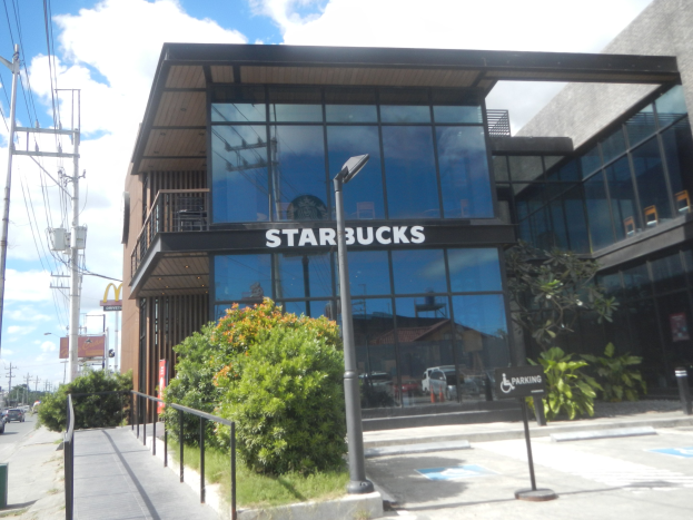 Außenansicht eines Starbucks-Cafés an einer Straßenecke mit umgebender städtischer Infrastruktur und Vegetation.