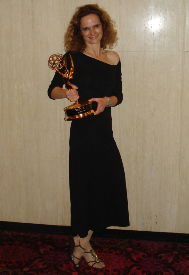 Eine Frau in einem schwarzen Kleid steht auf einem roten Teppich, lächelt und hält einen Emmy Award in der Hand, mit einer Wand im Hintergrund.