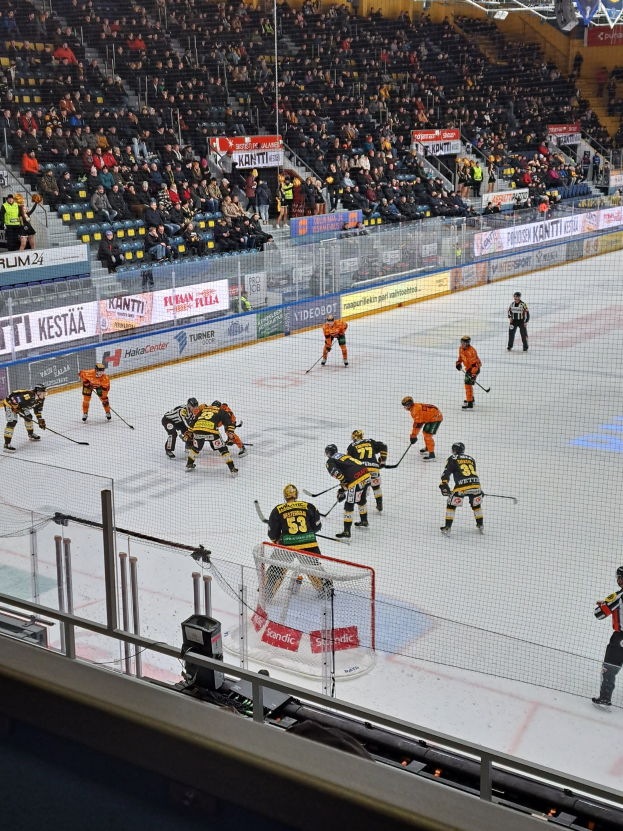 Eine Gruppe von Menschen, die Hockey auf einem Eisstadion spielen, umgeben von einem Zaun, mit Zuschauern im Hintergrund und beleuchteter Stadionbeleuchtung oben.