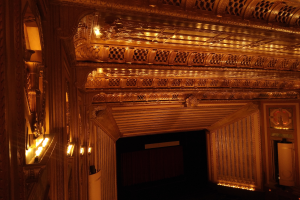 Das Innere eines Theaters mit dunklem Hintergrund, beleuchtet von Lampen links und rechts, mit einer kunstvoll gestalteten Decke.