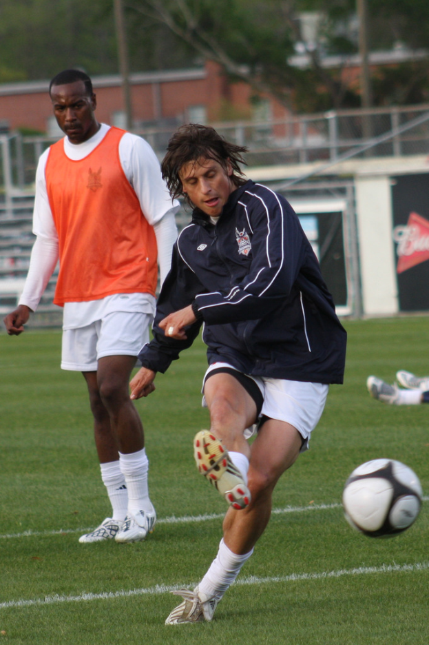 Ein Fußballspieler in einer schwarzen Jacke und weißen Shorts schießt einen Ball, während ein anderer Spieler in einer orangefarbenen Jacke hinter ihm steht und ein umzäunter Bereich mit Bäumen im Hintergrund zu sehen ist.