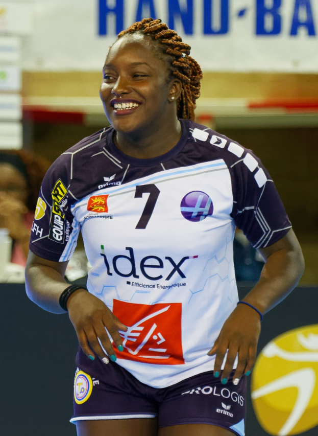Professionelle Handballspielerin in lila-weißer Uniform, die selbstbewusst auf einem Feld lächelt, mit "FIVB Handball-Weltmeisterschaft 2019"-Schilder im Hintergrund.