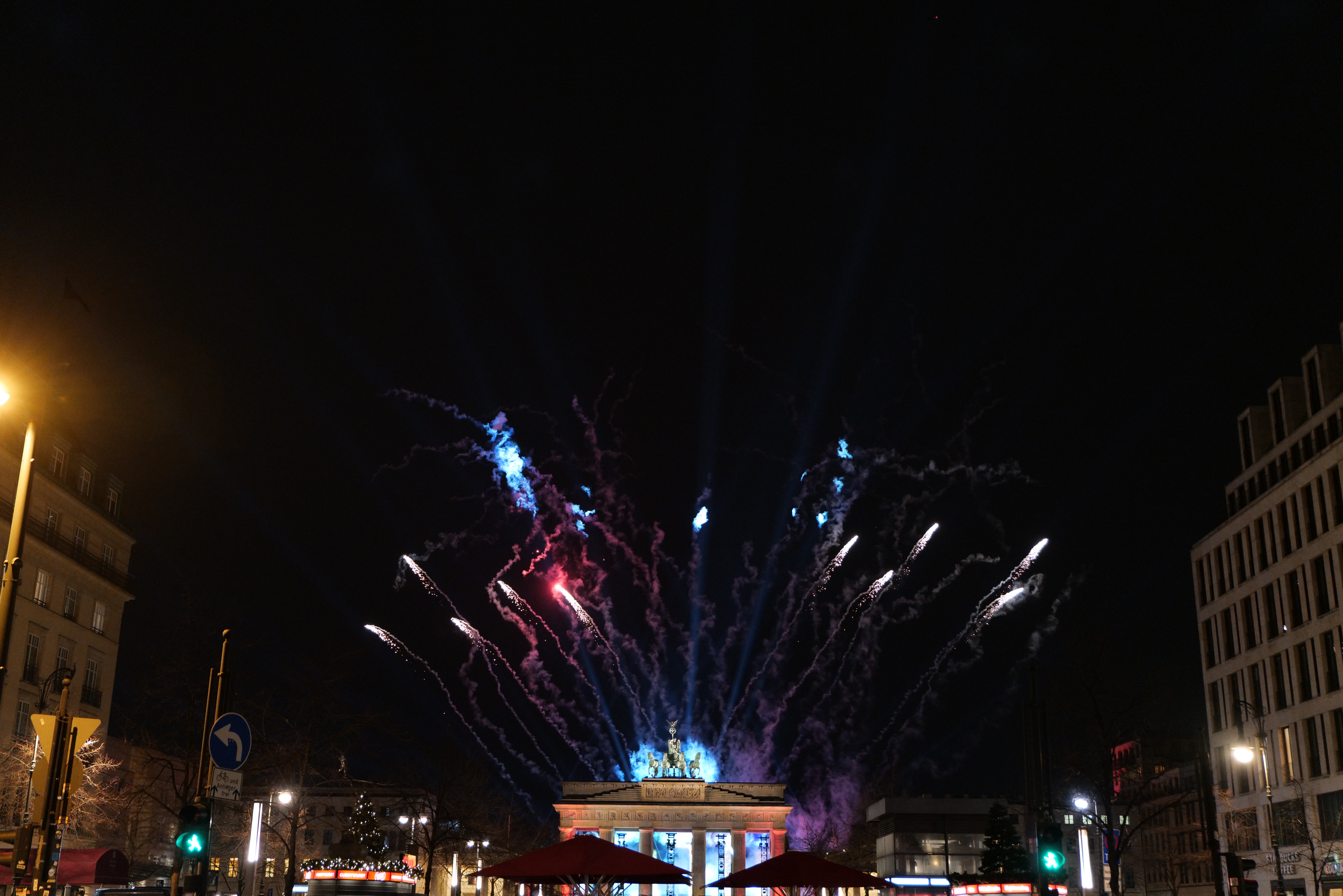 Eine belebte Straßenkreuzung in Berlin, Deutschland bei Nacht, beleuchtet von Straßenlaternen und farbenfrohen Feuerwerken am Himmel, mit Fahrzeugen, Fußgängern, Regenschirmen, Bäumen, Gebäuden, Schildern, Verkehrsampeln und einem Tor mit Säulen.