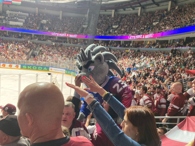 Eine Gruppe von Menschen steht vor einem Publikum bei einem Hockey-Spiel, mit einer Person in einem Kostüm umgeben von einem Metallzaun, einem Banner, Pfählen und einer Decke mit Deckenleuchten.