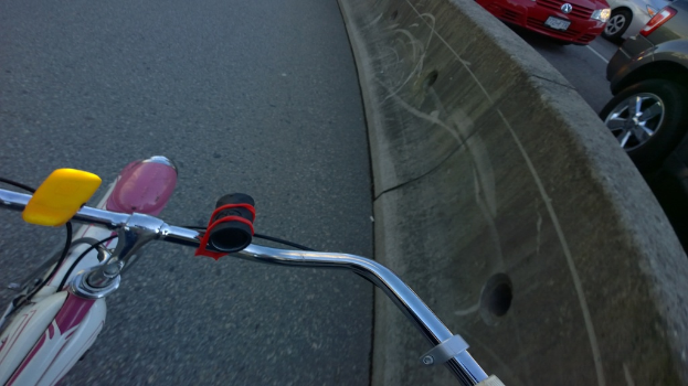 Ein Fahrradlenker befindet sich unten im Bild, mit Fahrzeugen, die rechts auf der Straße fahren, und einer Mittellinie.