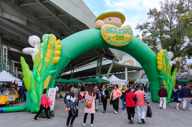 Eine Gruppe von Menschen geht auf einer Straße vor einem großen grünen Torbogen an der Taipei Expo Farmers Market vorbei, das mit einer Cartoon-Abbildung und Text verziert ist und von Zelten, Fahrzeugen und anderen Gegenständen umgeben ist, mit Gebäuden, Bäumen und einem klaren blauen Himmel im Hintergrund.