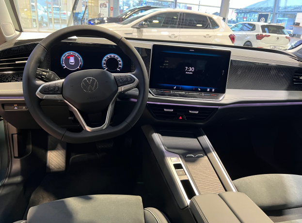 Das Innere eines Volkswagen e-Golf mit einem Lenkrad, Armaturenbrett, Bildschirmen, Knöpfen und Sitzen, mit einem Blick auf andere Autos, Gebäude, Bäume, Pfosten und den Himmel durch die Glastüren.