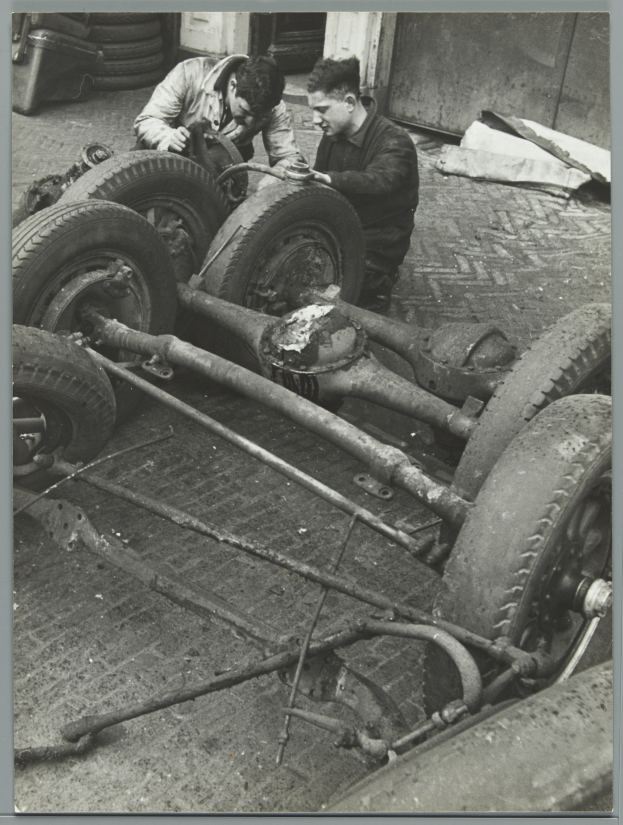 Zwei Männer arbeiten an einem Auto in einer Garage, halten Werkzeuge, umgeben von Reifen und anderen Gegenständen, auf einem Schwarz-Weiß-Bild.