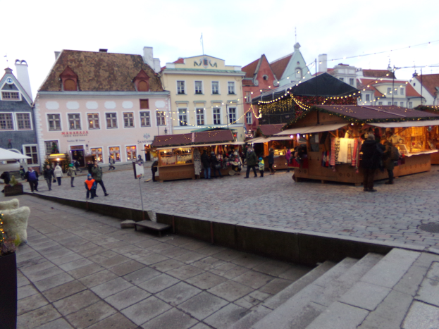 Ein geschäftiges Weihnachtsmarkt in Tallinn, Estland mit Menschen um dekorierte Stände, festliche Lichter, bewölktem Himmel, Treppen fürher und Topfpflanzen im Bereich.