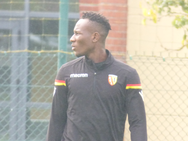 Ein Mann In Einer Schwarzen Jacke Steht Auf Einem Fussballfeld, Umgeben Von Einem Zaun Und Einem Baum, Und Sieht konzentriert Aus