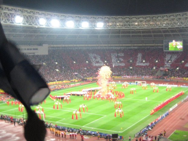 Ein Stadion mit Flutlicht, Eisengittern, Zuschauern, stehenden Personen, Werbetafeln, Anzeigetafeln, einer ├ťberwachungskamera und Fahnen und Deko.