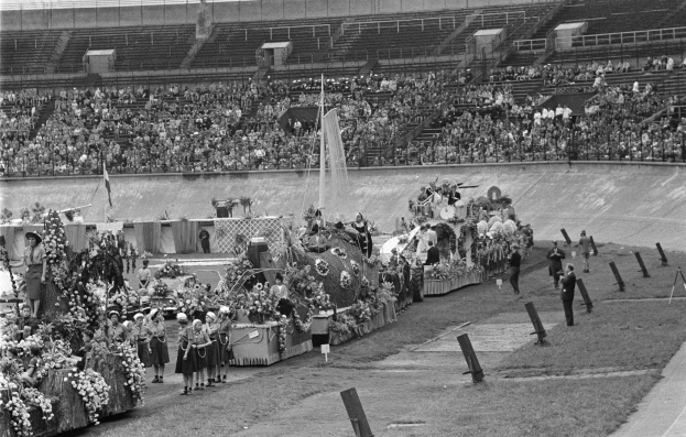 Schwarzes und weißes Foto eines Umzugs in einem Stadion mit Menschen, die stehen und sitzen, einem zentralen Springbrunnen und Blumensträußen auf Fahrzeugen.