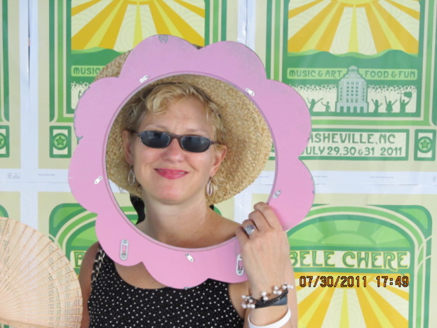 Eine Frau in einem cremefarbenen Hut, schwarzen Brillengläsern und einem schwarzen Kleid steht vor Plakaten.