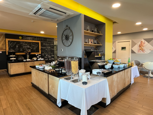Ein Restaurantinnenraum mit Tischen und Stühlen, Speisen, einer Tafel mit Text, einer Wanduhr, Deckenleuchten, einer Klimaanlage und einer Tür im Hintergrund, Located in the Hôtel de Ville in Paris.