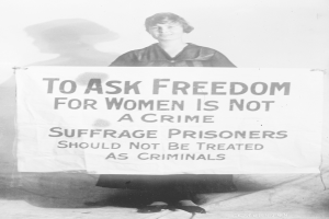 Schwarz-weiß-Fotografie einer Frau, die ein Transparent hält, auf dem steht: "Für die Freiheit der Frauen zu kämpfen ist kein Verbrechen, Suffragetten sollten nicht wie Kriminelle behandelt werden."