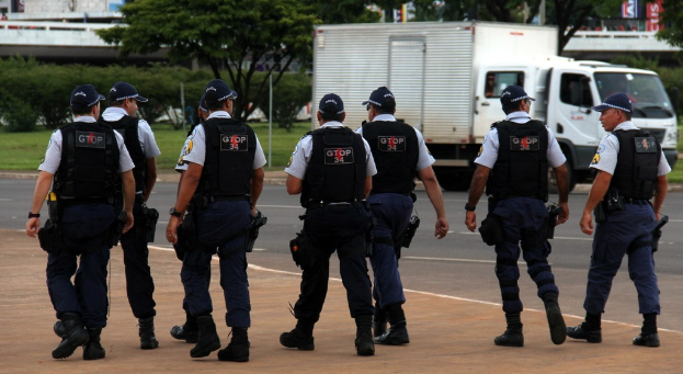 Eine Gruppe von Beamten in Uniformen, einschließlich Mötzen, Hemden, Schutzwesten, Uhren, Hosen und Schuhen, die auf einer Straße mit einem geparkten Lkw rechts, Gebäuden, Bäumen, Pflanzen, Masten, Bannern und einer Br√∞cke im Hintergrund gehen.