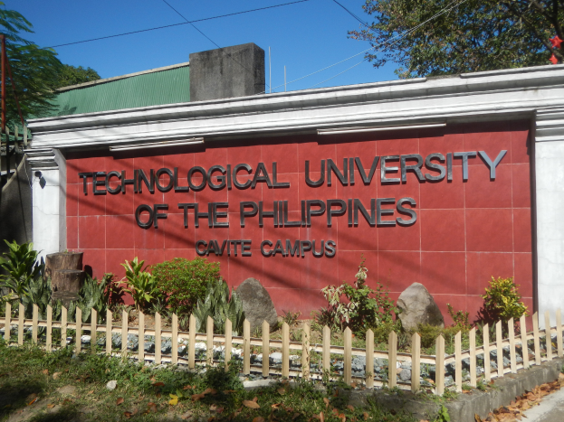Die Technologische Universität der Philippinen, Cavite Campus, mit einer Wand mit Text, umgeben von Pflanzen, Steinen, einem Holzzaun, Gras, trockenen Blättern, einer Gruppe von Bäumen, Pfählen, Drähten und einem bewölkten Himmel.